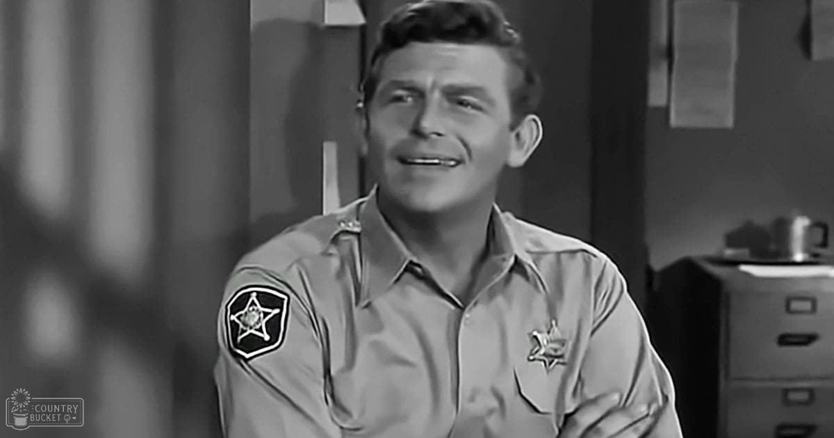 A young adult male in a relaxed, light gray or beige collared shirt with long sleeves and patches on an indoor office-like setting with a file cabinet and papers.