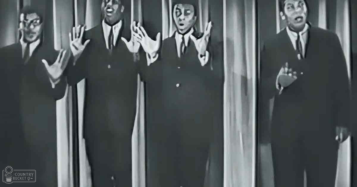 A group performance scene with four adult male performers in dark suits, set against a black-and-white theatrical backdrop.