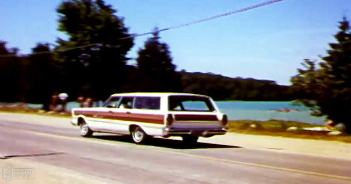 A vintage station wagon parked on an asphalt road near a lakeside, surrounded by trees in a rural area.