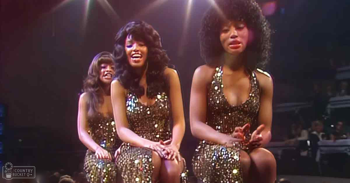 Adult females in sleeveless, sequined gold jumpsuits perform on a dimly lit stage at an evening event.