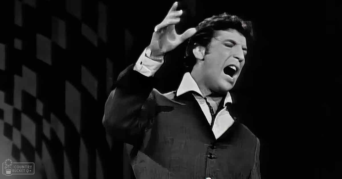 An adult male performer in a dark indoor stage setting with a checkered backdrop wearing a classic dark suit jacket over a white collared shirt.