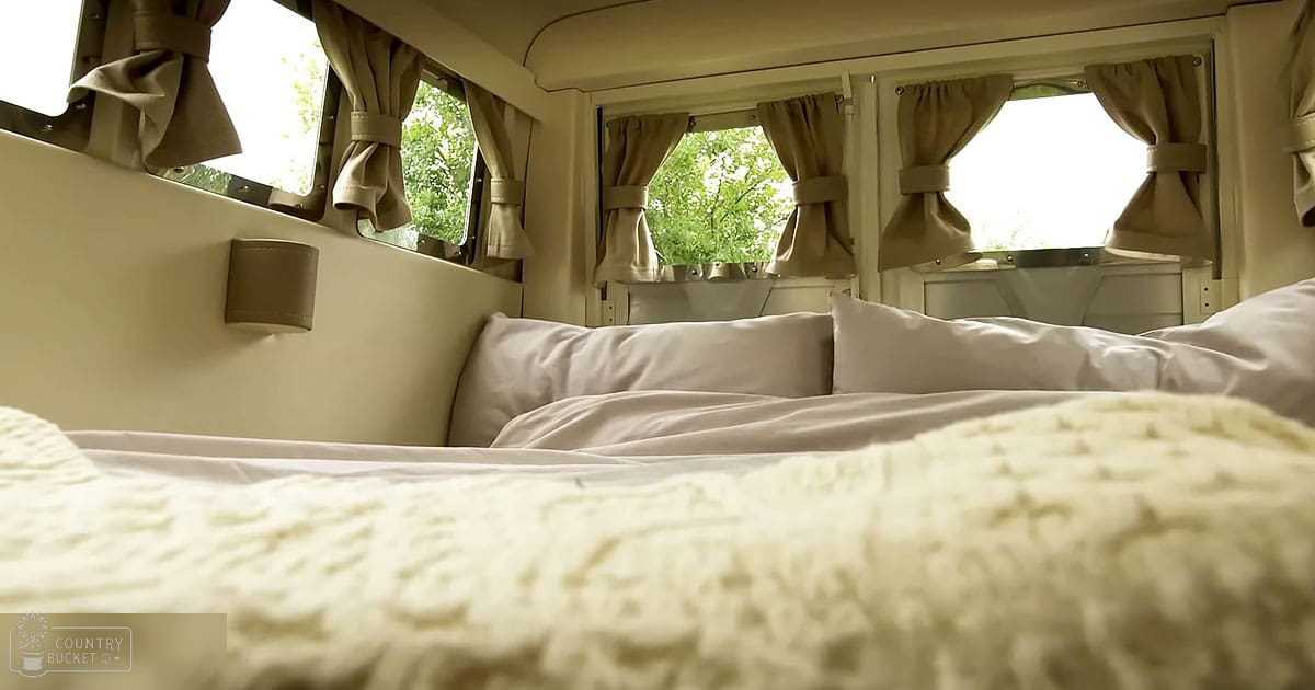Cozy interior space inside a camper van or similar vehicle, recreational area for travel; beige curtains, white bedding, and beige wall paneling create a comfortable setting with natural outdoor light and greenery visible through windows.