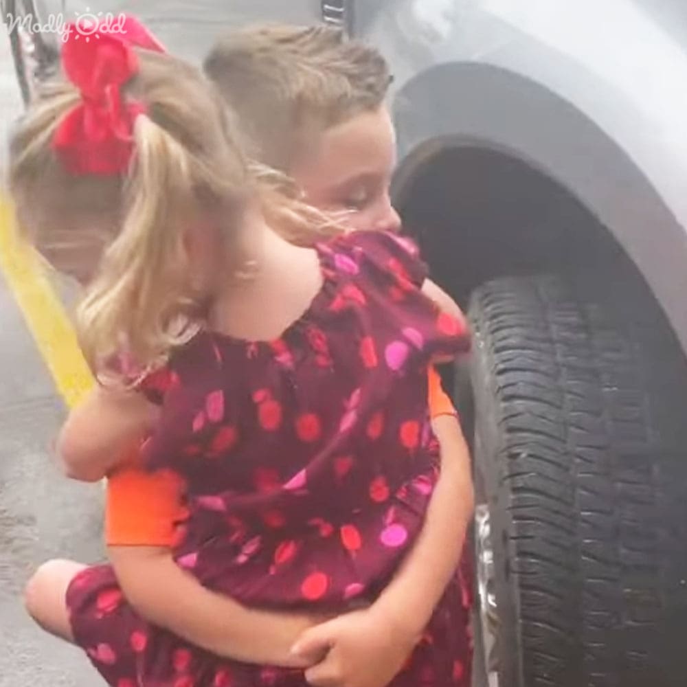 A young female child in a dark red polka dot dress held by another child wearing an orange top and pink polka dot skirt, near a vehicle tire on what appears to be a sunny day outdoors.