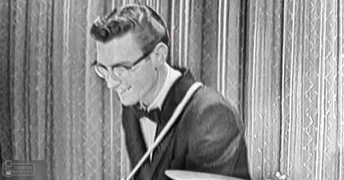 A young adult male playing drums indoors, wearing a dark suit jacket and white shirt with glasses.