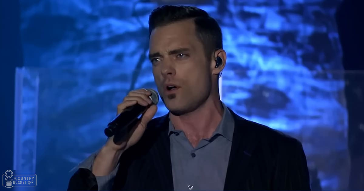 An adult male singer performing on stage, wearing a dark blazer over a light gray button-up shirt with long sleeves, holding a microphone against a blue-toned background with abstract patterns.