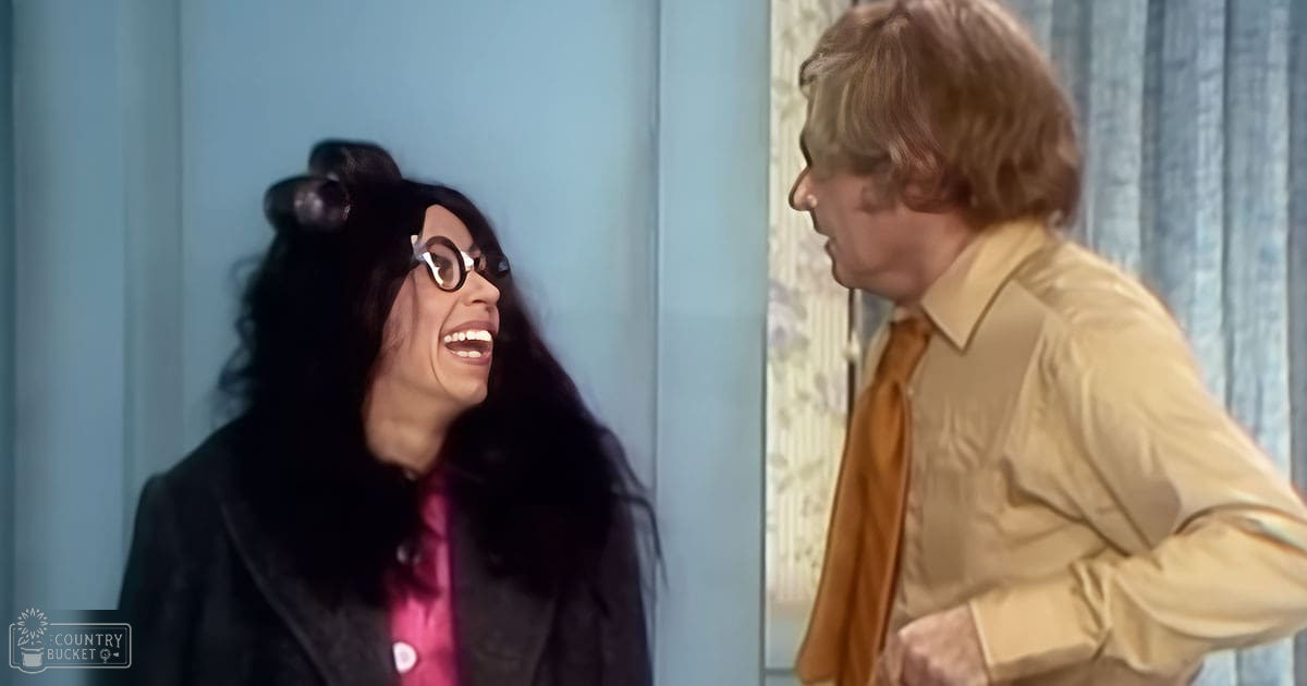 An adult female in a black blazer and pink shirt indoors with light blue walls and curtains, wearing round glasses.