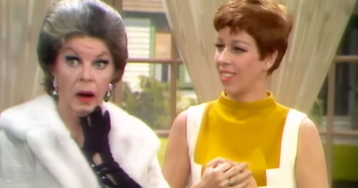 Indoor scene with two individuals, one older adult female wearing a white fur coat and another young adult female in a yellow panel top; light-colored curtains and green background object suggest daytime setting.