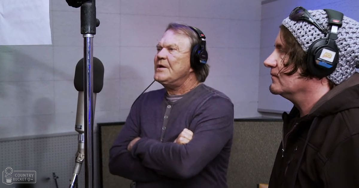 Middle-aged male in a recording studio wearing a dark gray button-up shirt with long sleeves and a crew neckline, surrounded by soundproofing panels typical of such environments.
