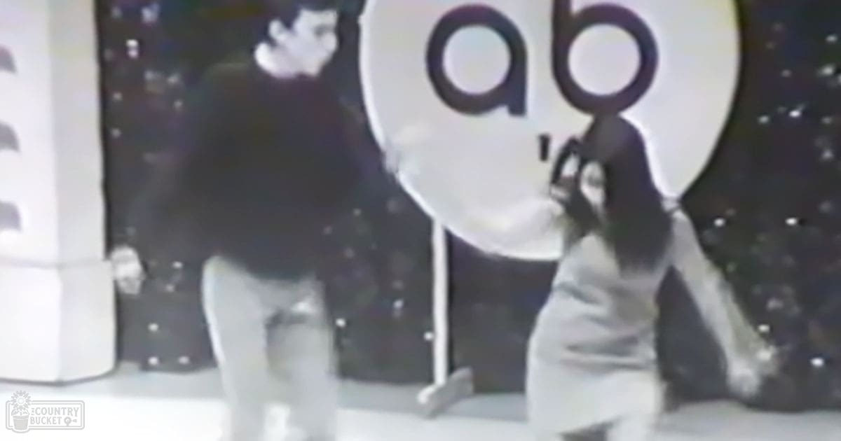 A young adult female wearing a loose-fitting, long-sleeved white dress stands in front of an educational setting with a large 'ab' sign and starry background. The indoor environment suggests a classroom or children's program space.