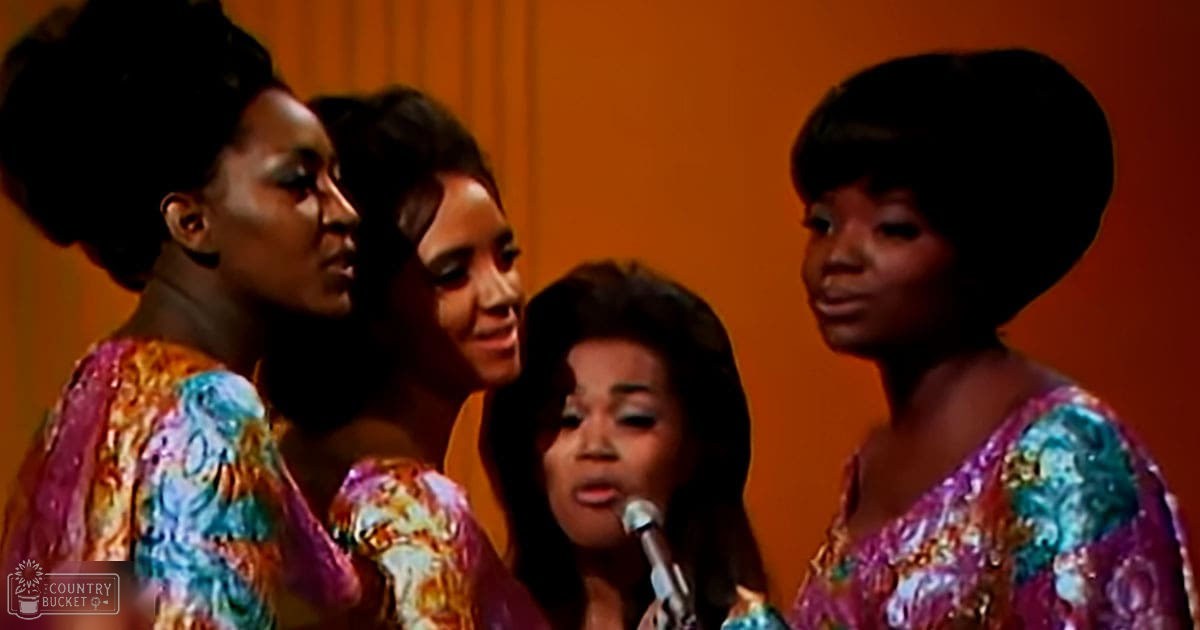 A group of adult female singers in a studio setting with vibrant patterned tops and an orange backdrop.