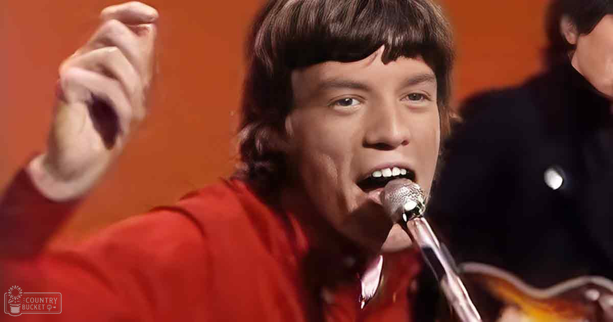 A young adult male singer in a red 1960s-70s era shirt on stage, with an adult male background figure wearing dark clothing and a white buttoned sleeve. The setting is brightly lit with orange backdrop.