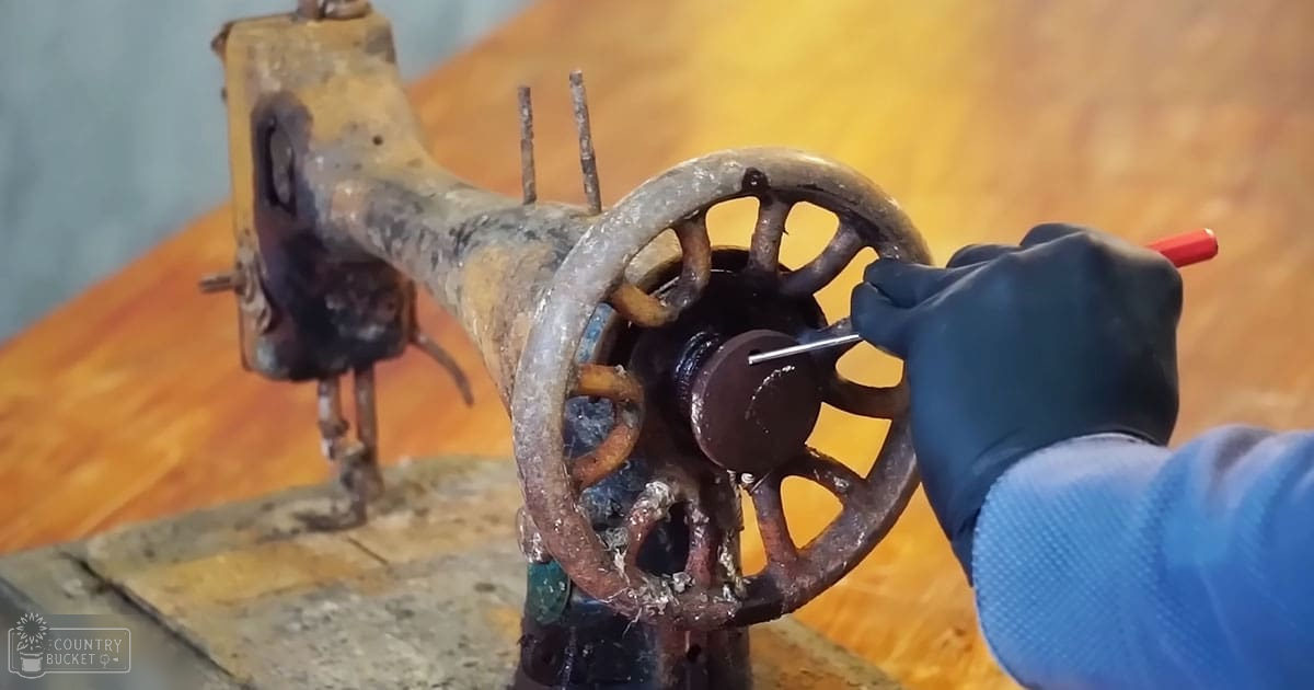 A gloved hand adjusts an old, rusted sewing machine with a hand crank in a workshop setting.