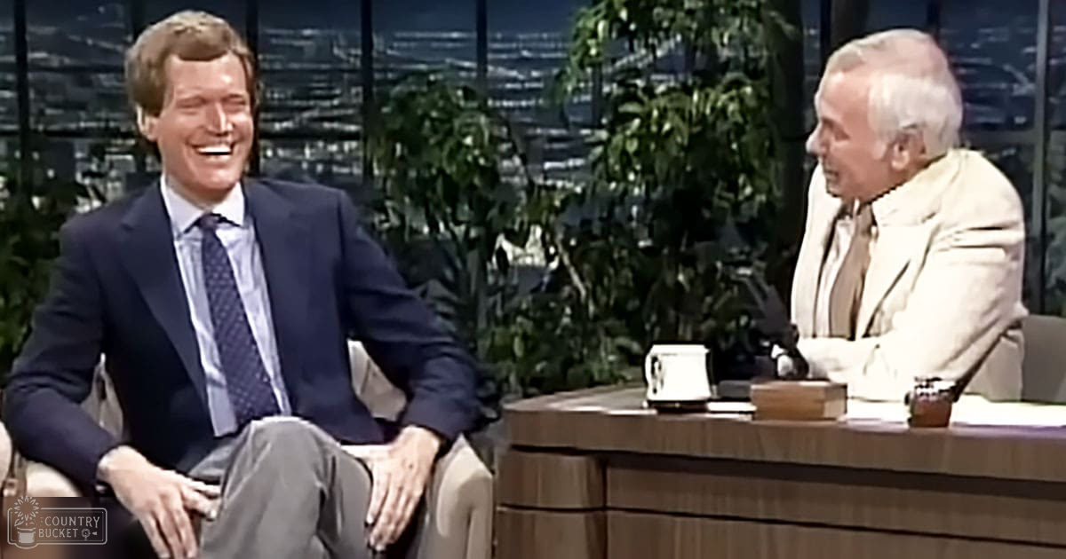 Adult male talk show host wearing a dark blue suit with patterned tie, seated at a desk with studio furniture and green plants in an indoor television studio setting.