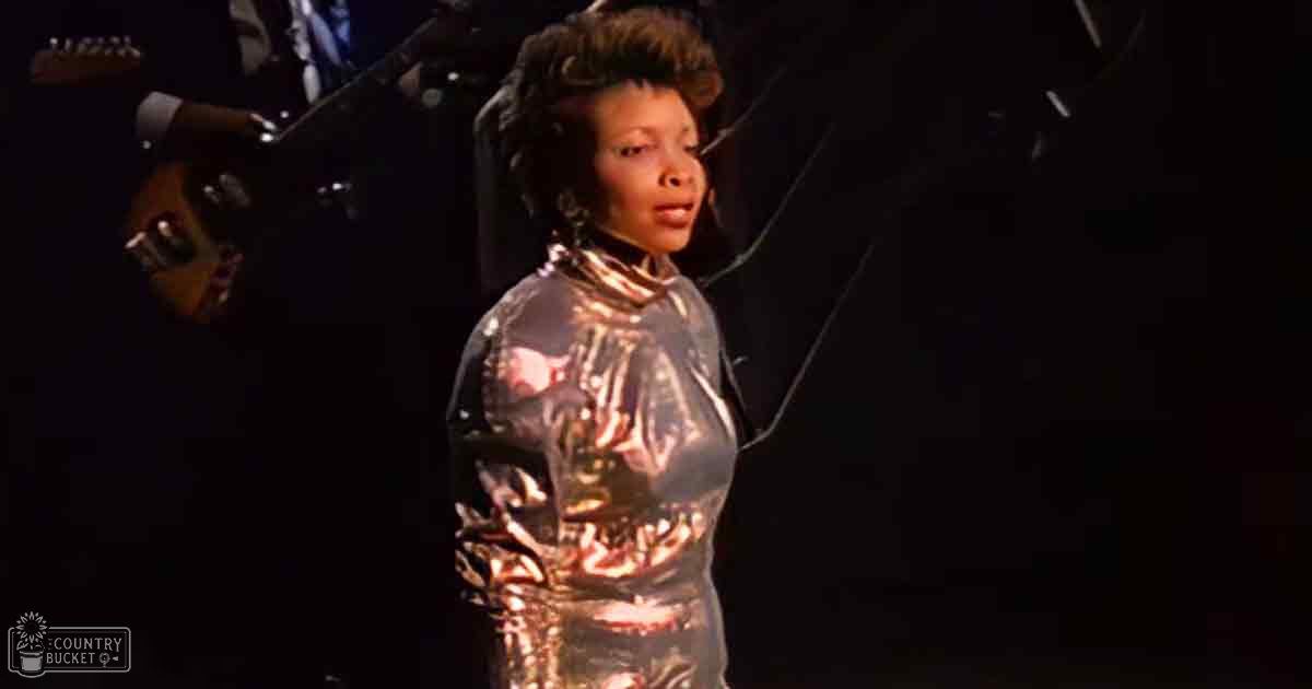 A female adult performer in a shiny, metallic silver outfit on stage at an indoor concert with dim lighting and a microphone stand visible behind her.