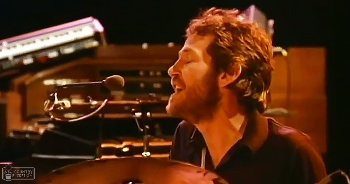 An adult male performer in a dark-colored shirt, performing on stage at an indoor concert venue with a drum set and amplifiers visible.
