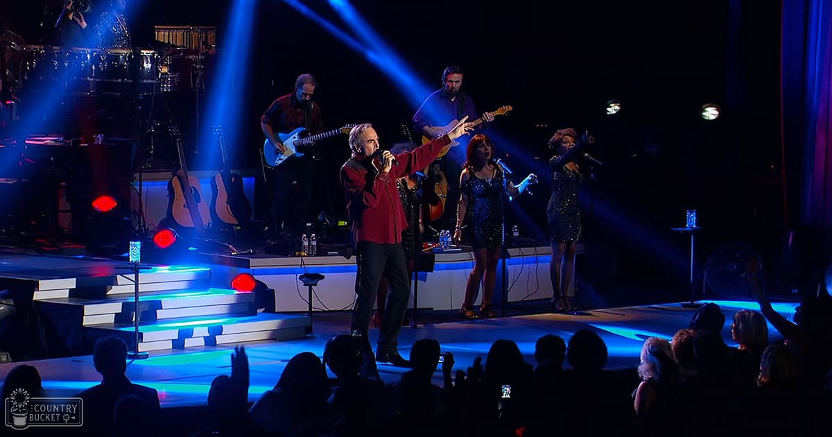 Adult male singer wearing a red jacket over a dark shirt and black pants on stage with two female singers in sparkly dresses at an indoor concert hall; blue stage lighting, musical instruments present.