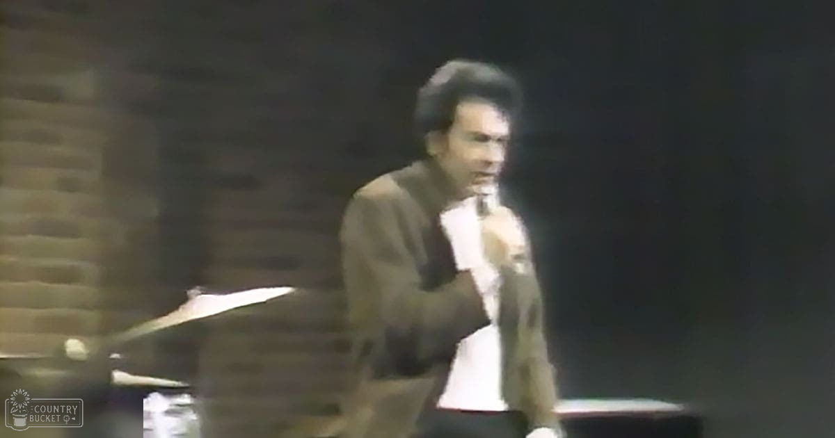 An adult male singer in a dark blazer over a crisp white shirt, performing on stage with sheet music visible on a piano stand.