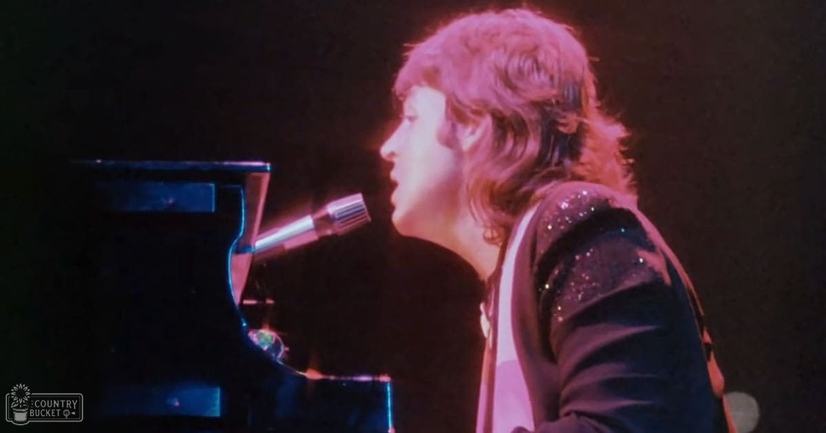 A male adult in a dark, sequined stage costume with full-length sleeves performs on an indoor concert stage with a grand piano and microphone visible.