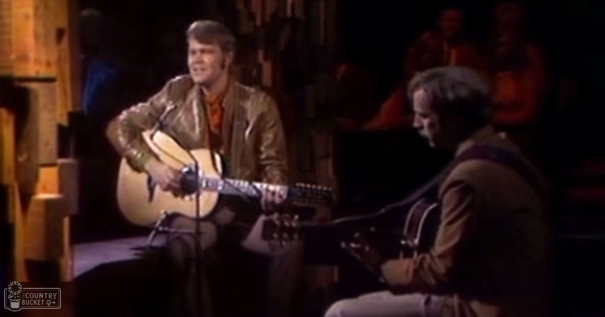 A male musician playing an acoustic guitar on a dimly lit stage wearing a brown leather jacket and red shirt; another adult male in a dark suit jacket also present.
