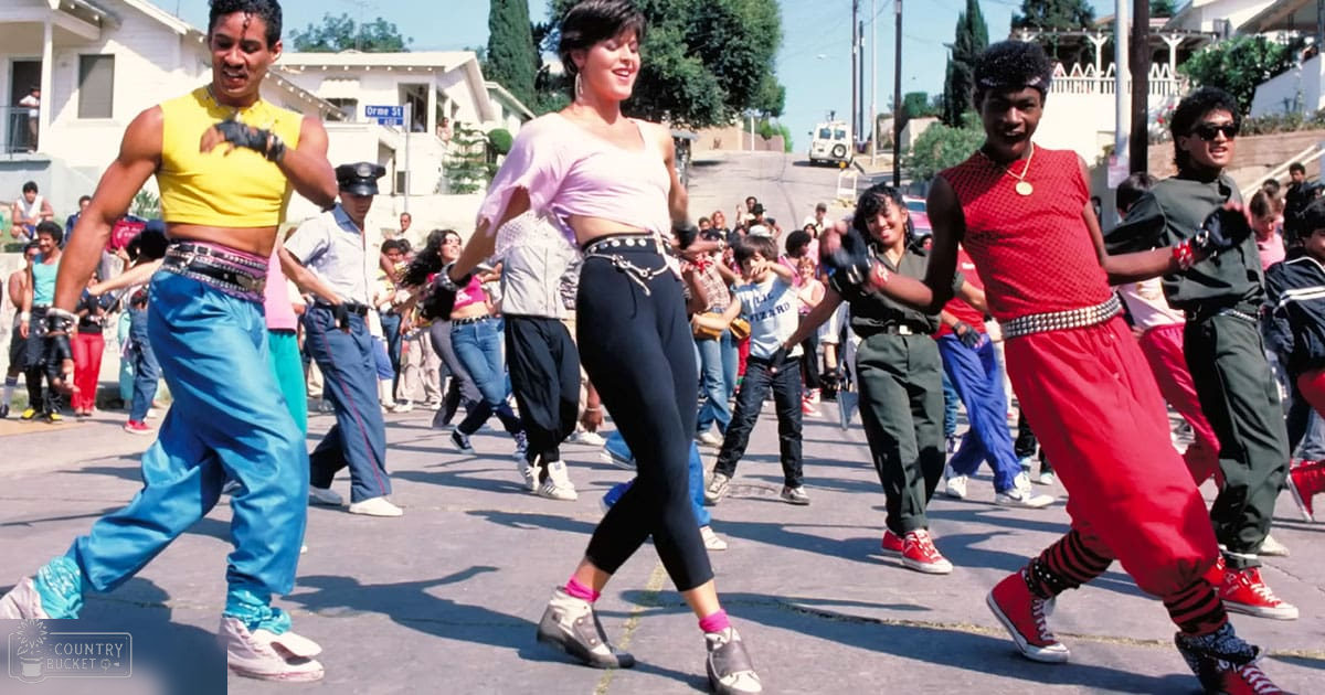 A group of young adults dancing on an urban street under a sunny sky, with various individuals in colorful clothing and accessories.