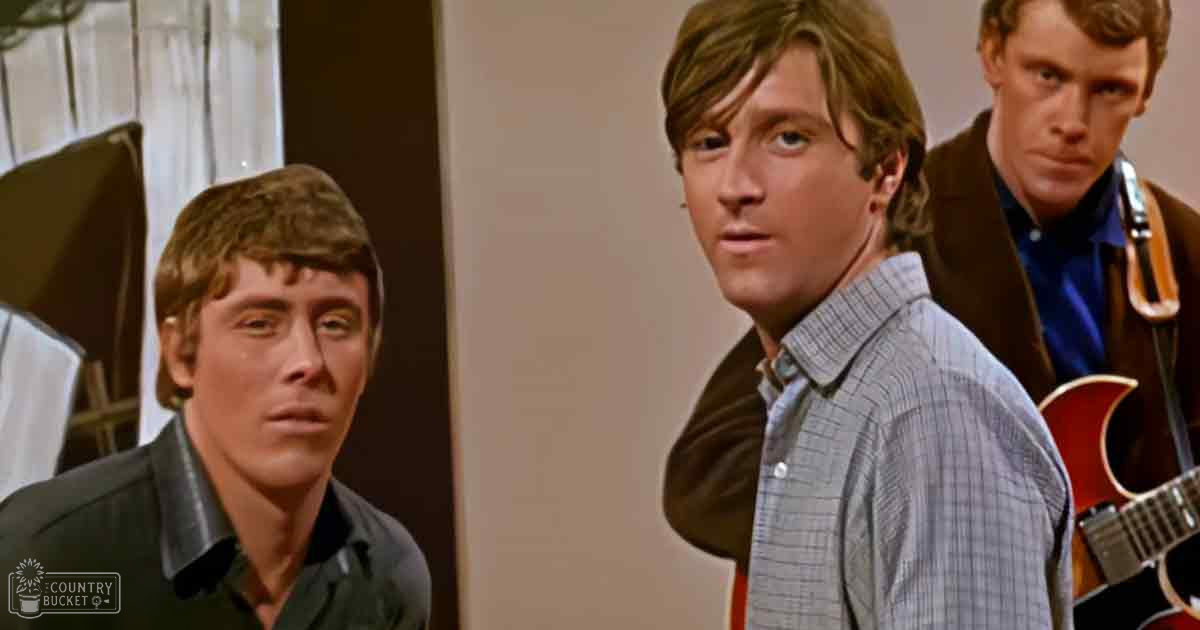 Three young adult males in a relaxed indoor setting, wearing casual shirts and jackets from the 1960s-1970s. Neutral-colored walls and curtains suggest daytime lighting.