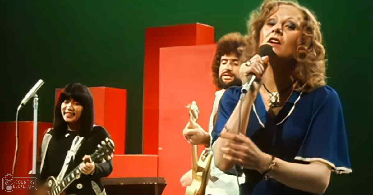 A middle-aged woman singing into a microphone, wearing a blue top with white trim and cotton-like material. She is accessorized with bracelets on her wrists.