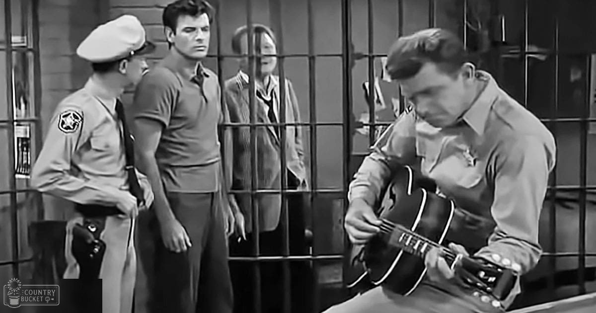 An adult male playing guitar with an officer nearby in a jail setting, black and white image.