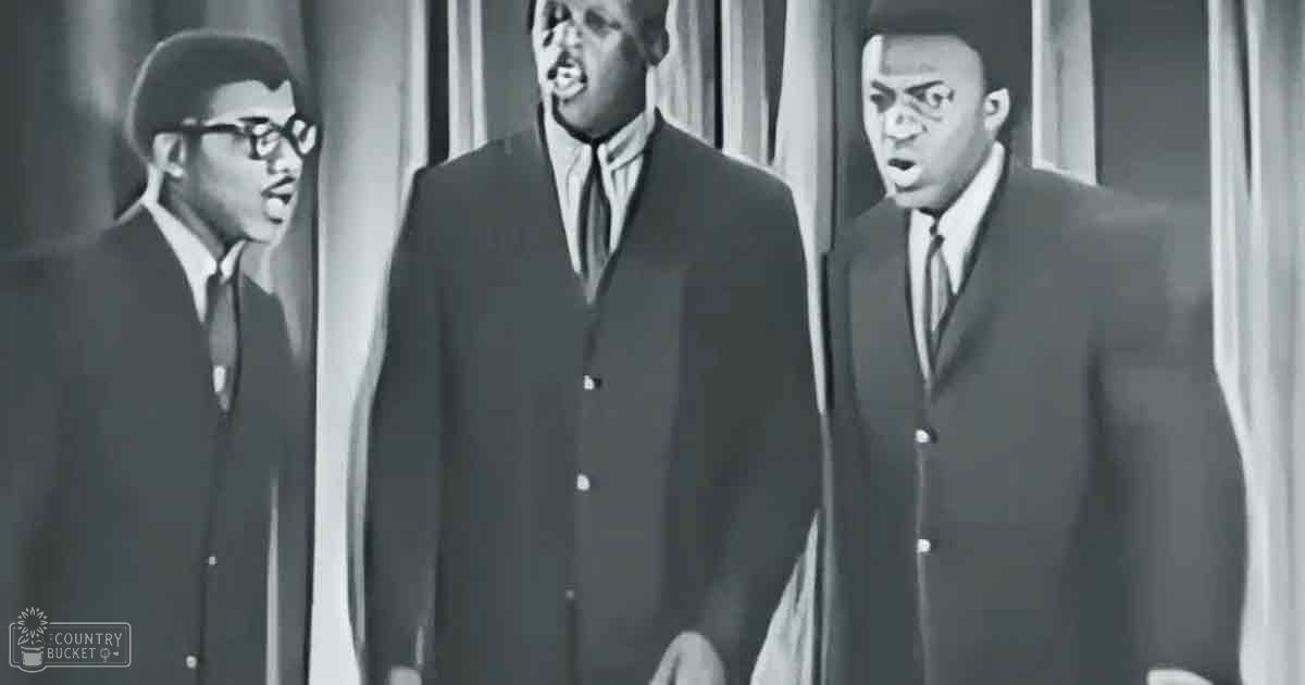 A musical performance scene with adults in their 30s or 40s wearing formal attire, set against vertical striped curtains suggesting a professional stage environment.