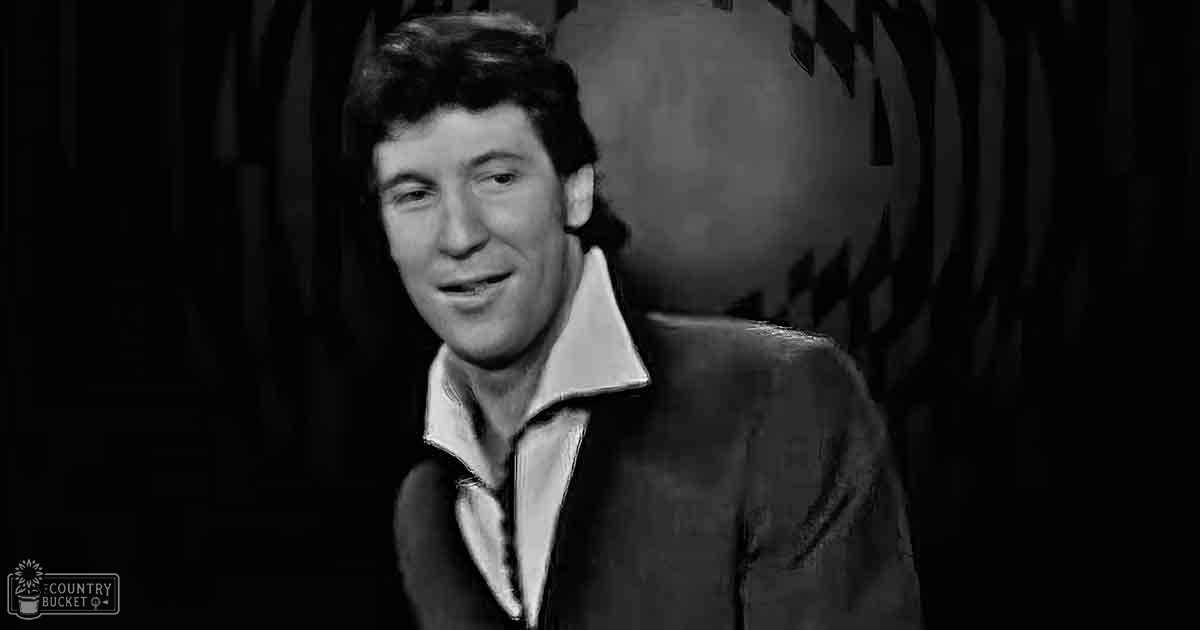 A middle-aged male wearing a dark textured jacket over a collared white shirt stands against an indoor backdrop with abstract foliage patterns, possibly in a studio setting.