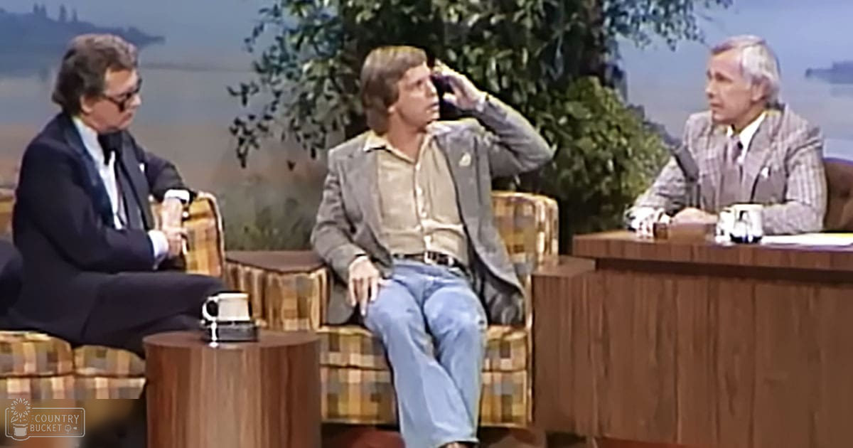 A young adult male in a light blue jeans and grayish-green jacket sits on a plaid chair, surrounded by two older adults seated behind a wooden desk, with an outdoor scene of trees and hills under natural daylight in the background.