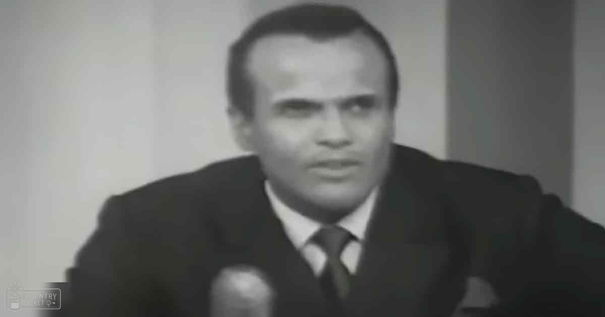 A middle-aged adult male in a tailored dark suit jacket, white shirt, and dark tie stands indoors with a microphone visible, suggesting a broadcast or recording session.