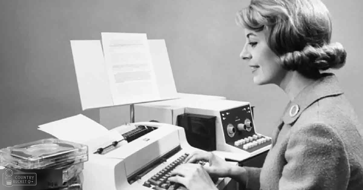 A woman wearing a tailored mid-20th century jacket works at an old computer terminal in an indoor office setting with stacks of paper documents.