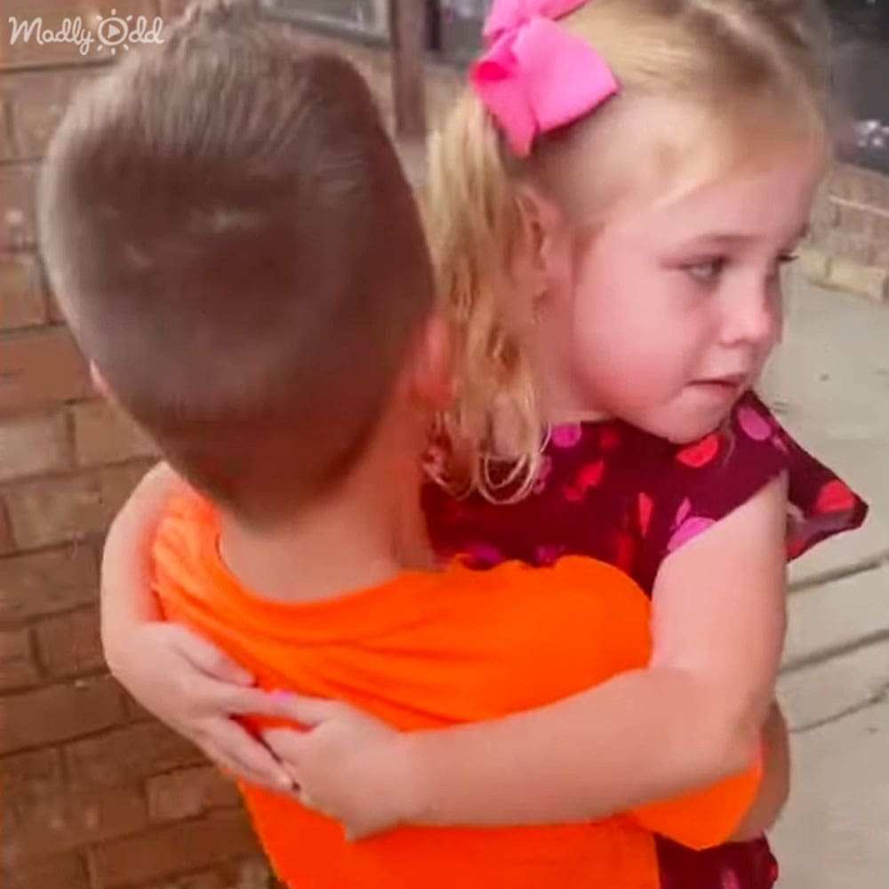 A toddler in a red polka dot dress with ruffled sleeves, held by an older child wearing an orange t-shirt near a brick wall on a sunny day.