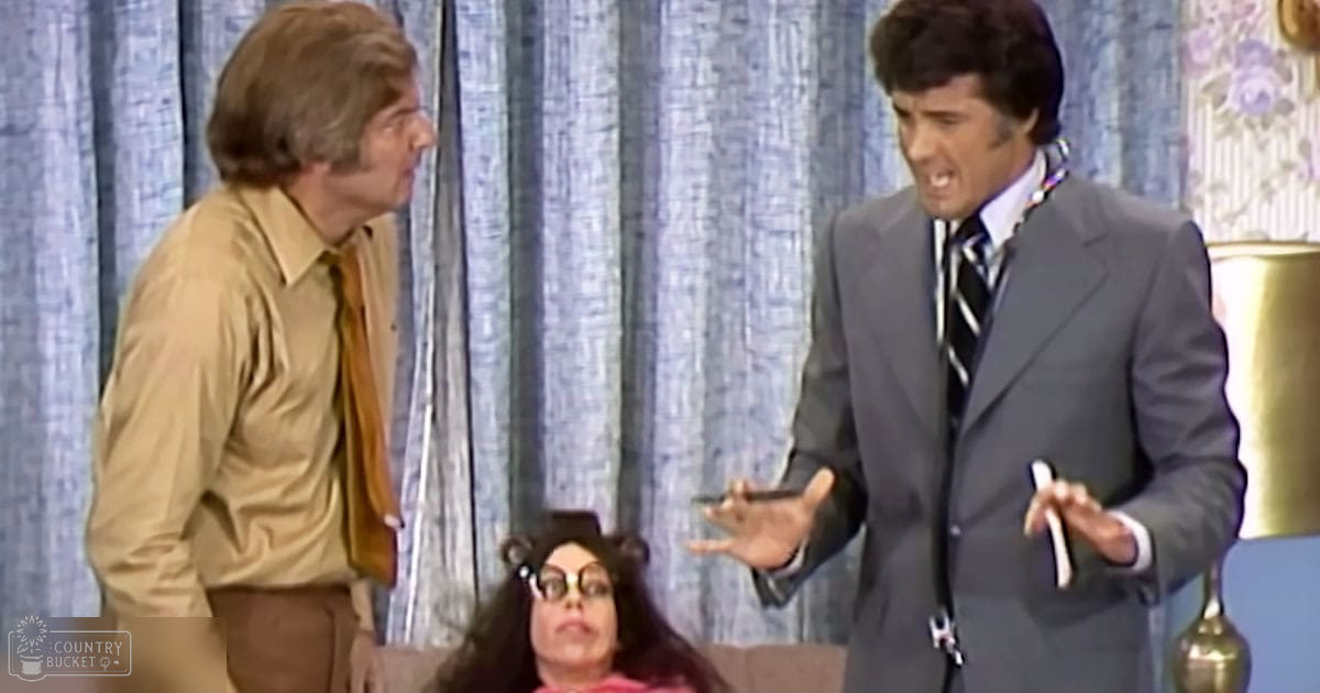 A comedic television scene set in the late 1960s or early 1970s with two adult males and one adult female, dressed in period-appropriate attire, interacting indoors against a blue curtain backdrop.