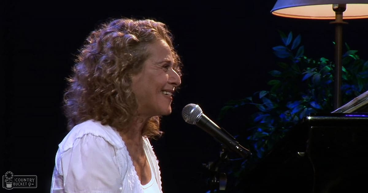 A middle-aged adult female in a modern casual white blouse with ruffled sleeves performs on stage under artificial lighting, surrounded by musical instruments and indoor decor.