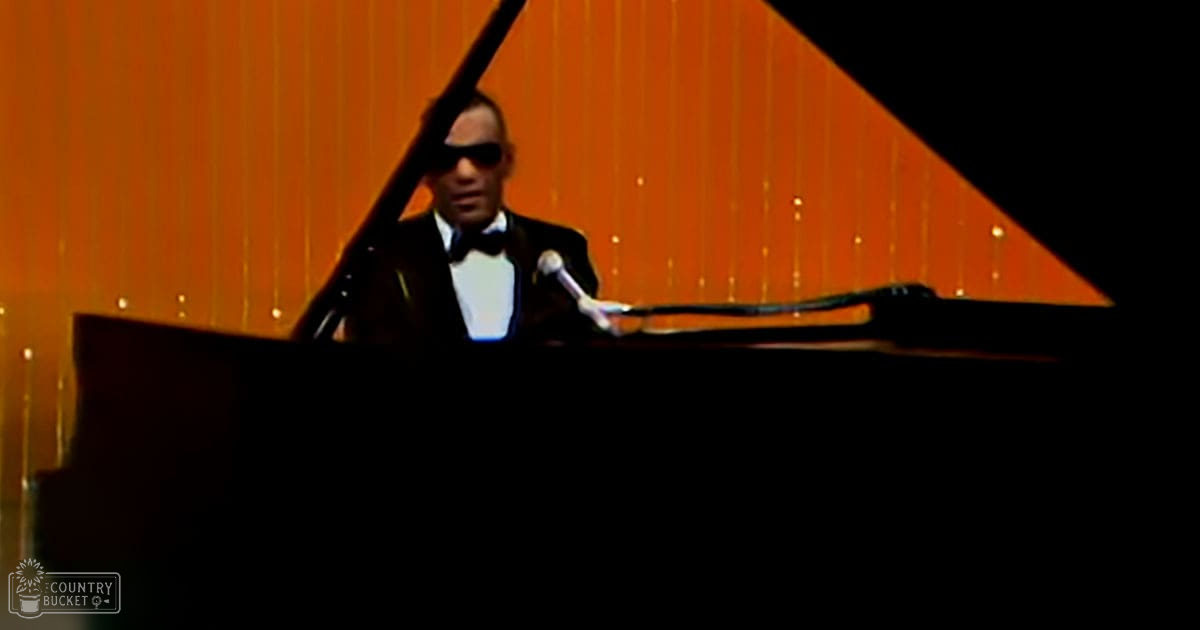 An adult male performer in a black tuxedo with white shirt and dark-rimmed sunglasses stands at a grand piano, set against vertical orange paneling under artificial stage lighting.