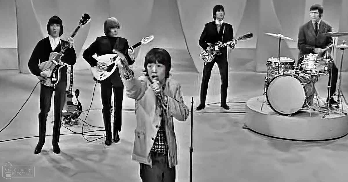 A young adult male singer wearing a light-colored jacket with a plaid shirt underneath, surrounded by band members in dark suits and ties, in an indoor music studio or performance space from the mid-20th century.
