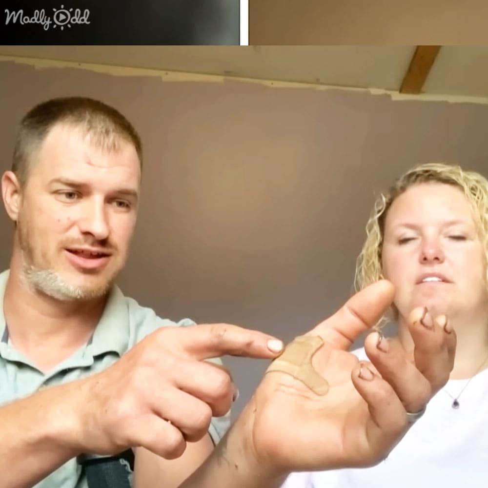 Two adults, a man in an indoor setting with unfinished ceiling and exposed beams, wearing a light green collared shirt; a woman holding her hand up with fingers slightly curled.