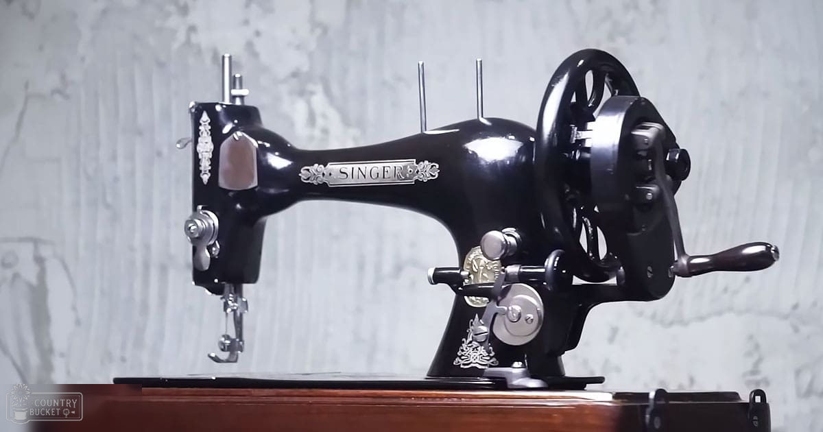 A vintage Singer sewing machine with ornate detailing, placed on a wooden base with a hand-crank mechanism, set against a textured concrete-like wall indoors.