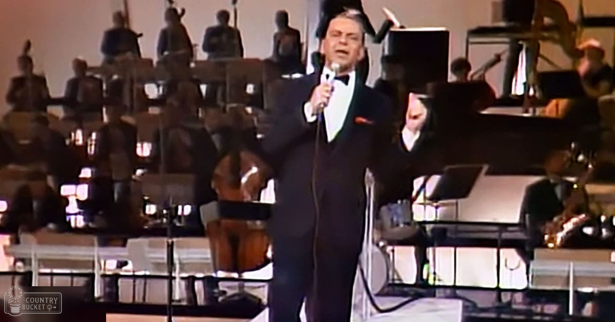 A middle-aged to older adult male performer in a dark tuxedo with white shirt and black bow tie, standing on stage before an orchestra in a mid-20th century concert hall setting. The background is illuminated by stage lighting, highlighting the performer while the orchestra remains in shadow.