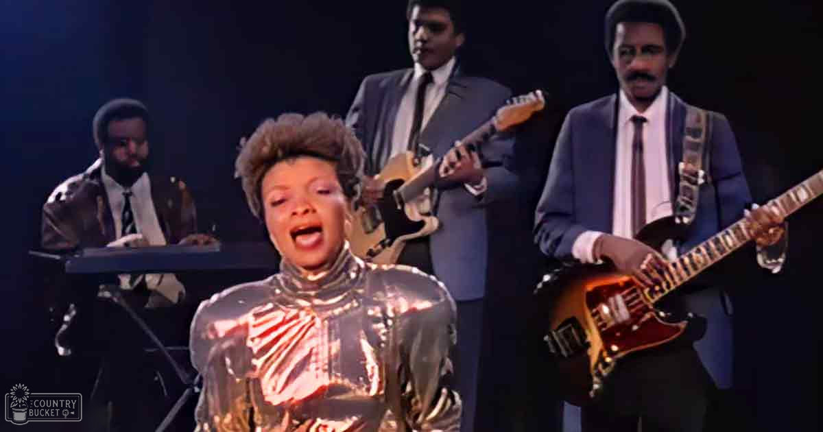 A female performer in a shiny, metallic 1970s-style outfit with long sleeves stands at center stage amidst her band dressed in coordinated dark suits under dim lighting.
