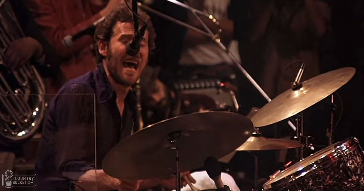 An adult male drummer performing on stage with a dark-colored shirt in an indoor music setting.