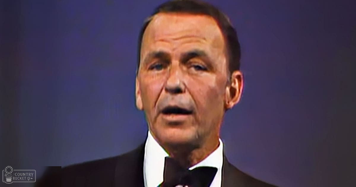 An older adult male wearing a classic tuxedo and bow tie, standing against a dark backdrop under stage lighting.