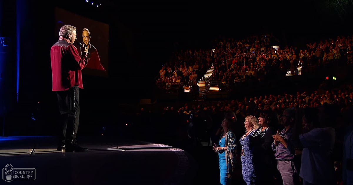Older adult male singer speaking on stage in a concert hall with an audience, wearing a red jacket and dark pants, surrounded by middle-aged female spectators dressed in patterned dresses under dim indoor lighting.