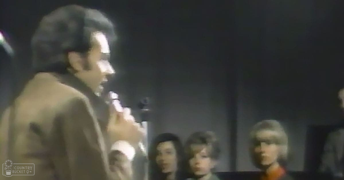 A man speaking into a microphone, possibly on stage, with two children in the background; dimly lit indoor setting, muted tones.