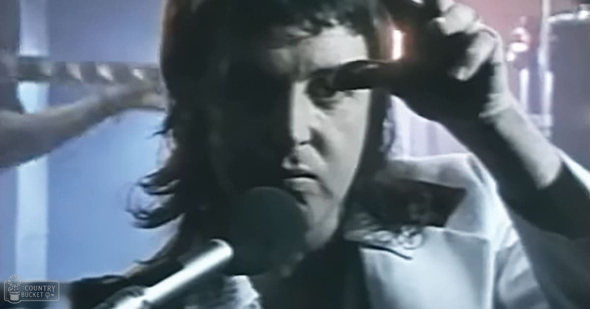 A young adult male singer wearing a white shirt with floral or paisley patterns performs on stage in what appears to be a music venue, with stage lighting and musical instruments visible in the background.