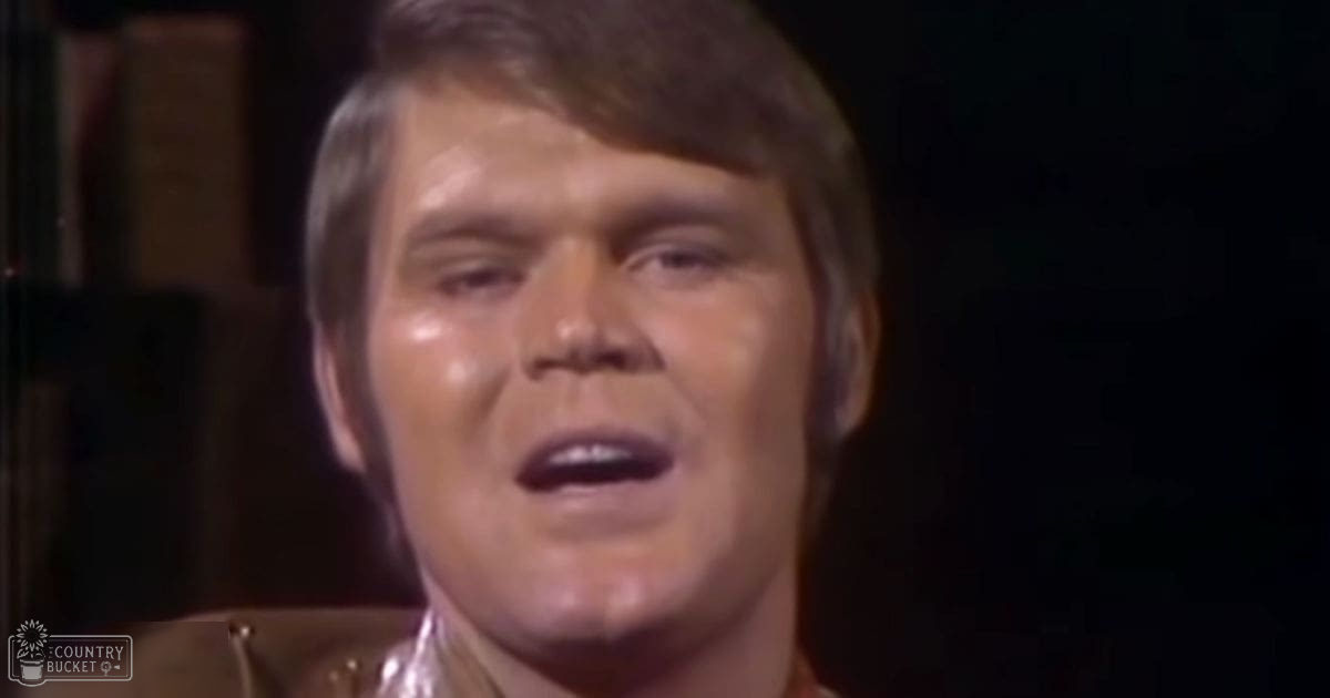 Middle-aged adult male wearing a form-fitting collared shirt with a reflective metallic finish stands indoors against a dark background, possibly stage or performance area.