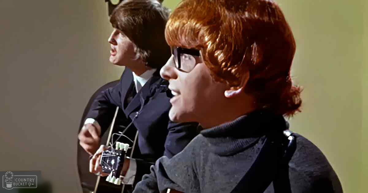 Young adult male wearing glasses and a dark turtleneck sweater indoors, holding an electronic device; another young adult male in the background wears a dark suit jacket with white shirt.