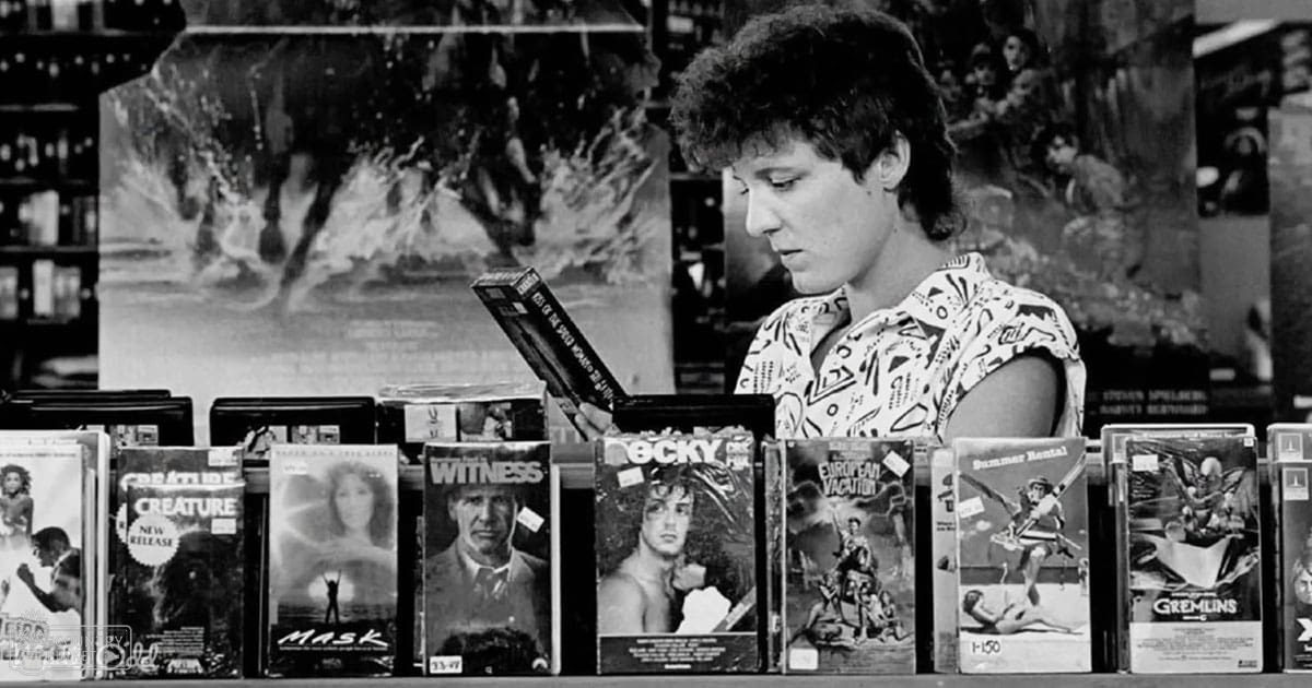 A young adult male in a video rental store wearing a white shirt with black patterns, surrounded by rows of VHS tapes and posters.