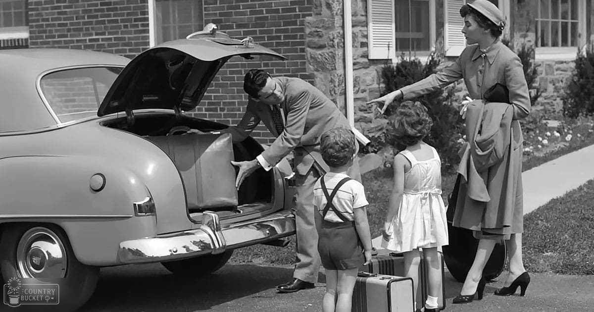 A family packing luggage into a vintage car in front of their suburban home on what appears to be a sunny day with lush greenery, likely spring or summer.
